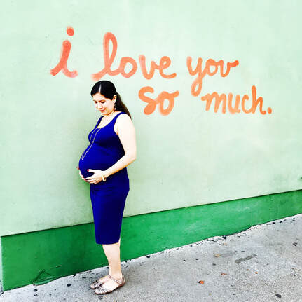 pregnant woman with brown hair and blue dress, in front of mural that reads 
